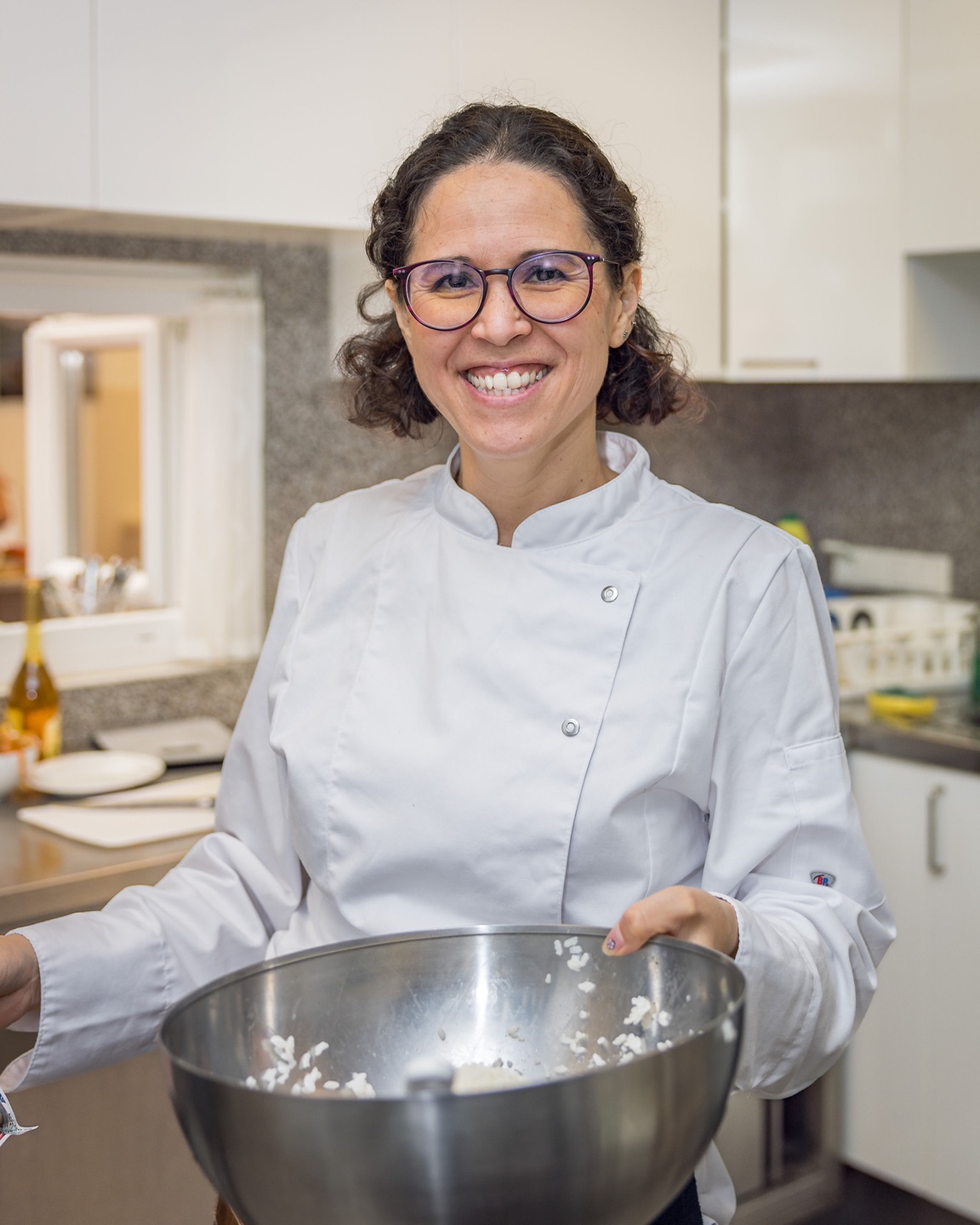 Ophelia Mariko Mahana smiling in a chef's jacket, holding a steel bowl of sushi rice in the kitchen